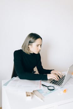Woman in black sweater working on a laptop surrounded by papers on a desk.
