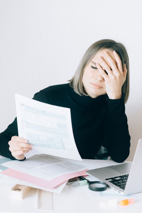 Free A woman in a black top sits at a desk stressed while reviewing documents, with a laptop nearby. Stock Photo