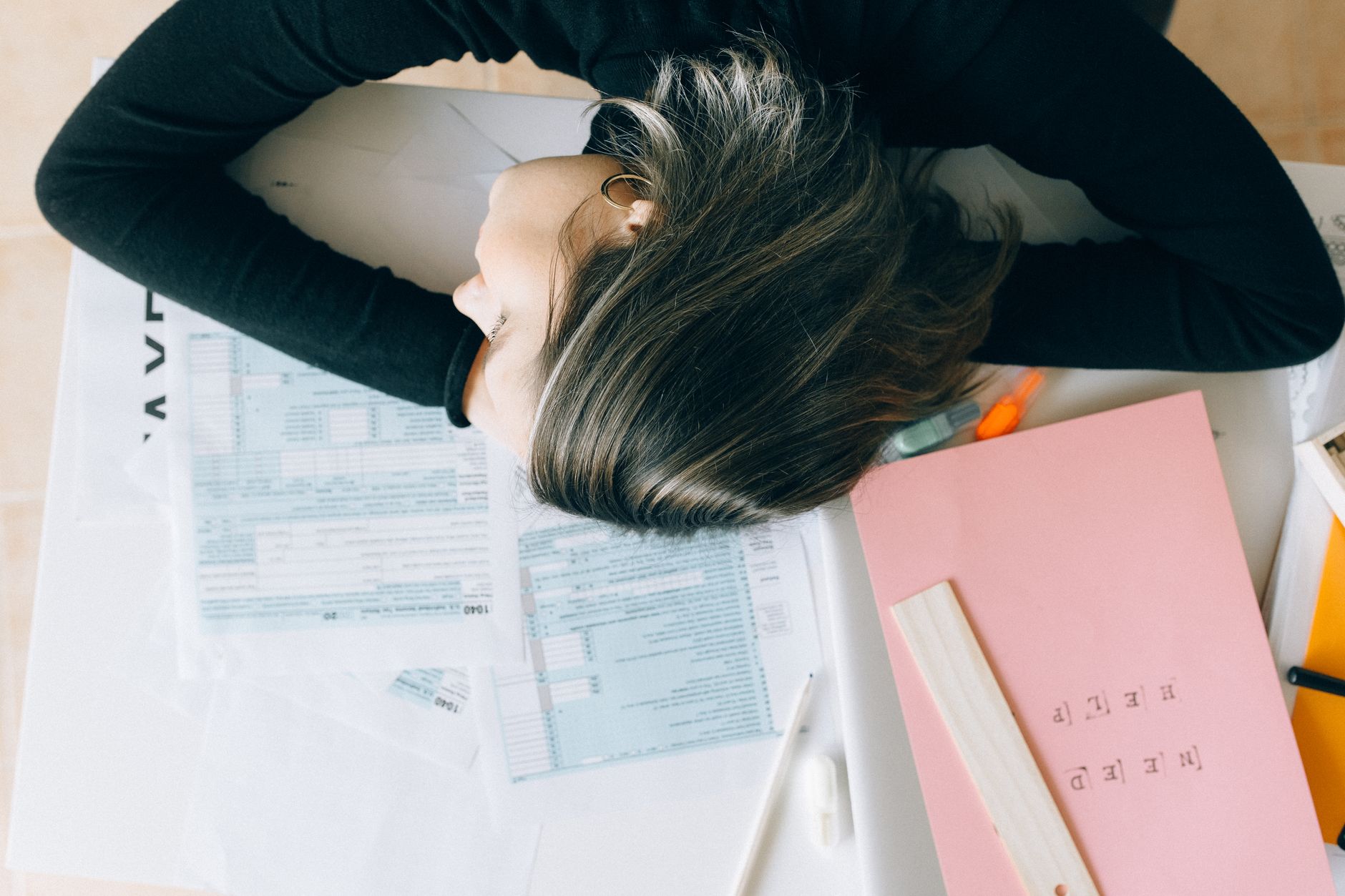 Exhausted woman resting her head on a desk cluttered with papers and a ruler, showing workplace fatigue.