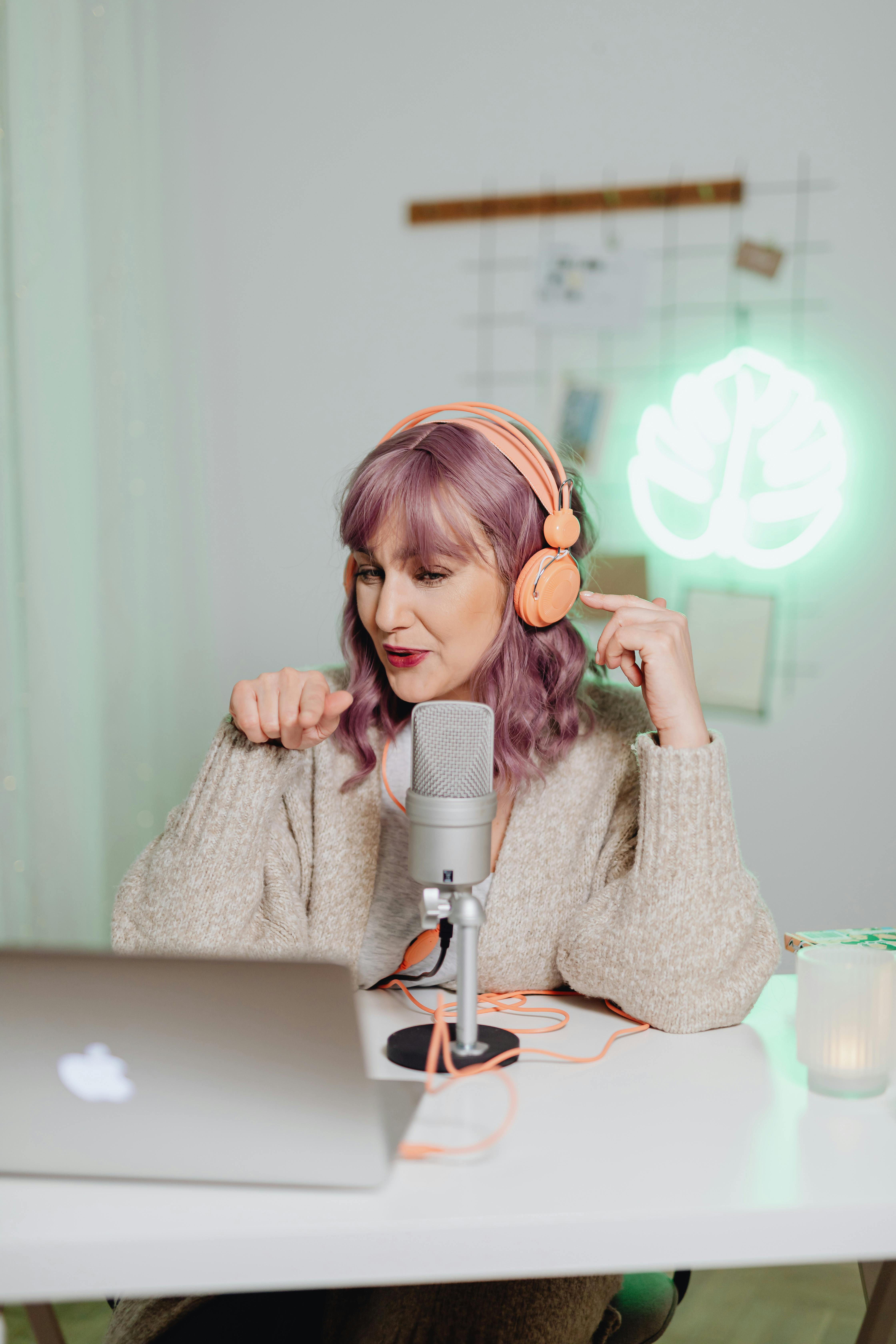 A woman recording a podcast indoors with a laptop and microphone, wearing headphones.