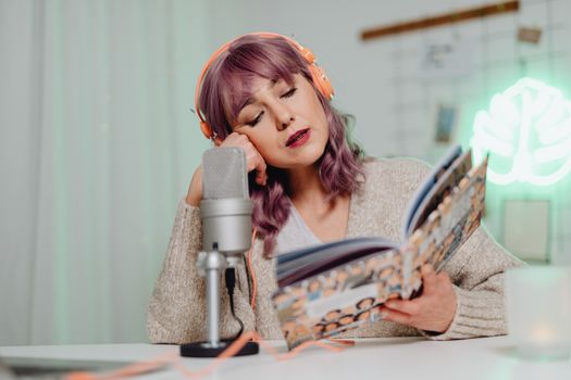 A woman wearing headphones records a podcast while reading from a book indoors.