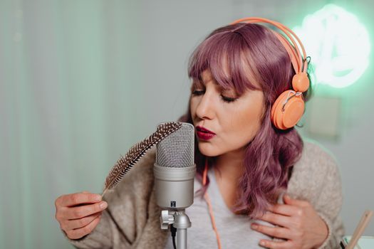 Woman with purple hair and headphones doing ASMR with a microphone and feather indoors.