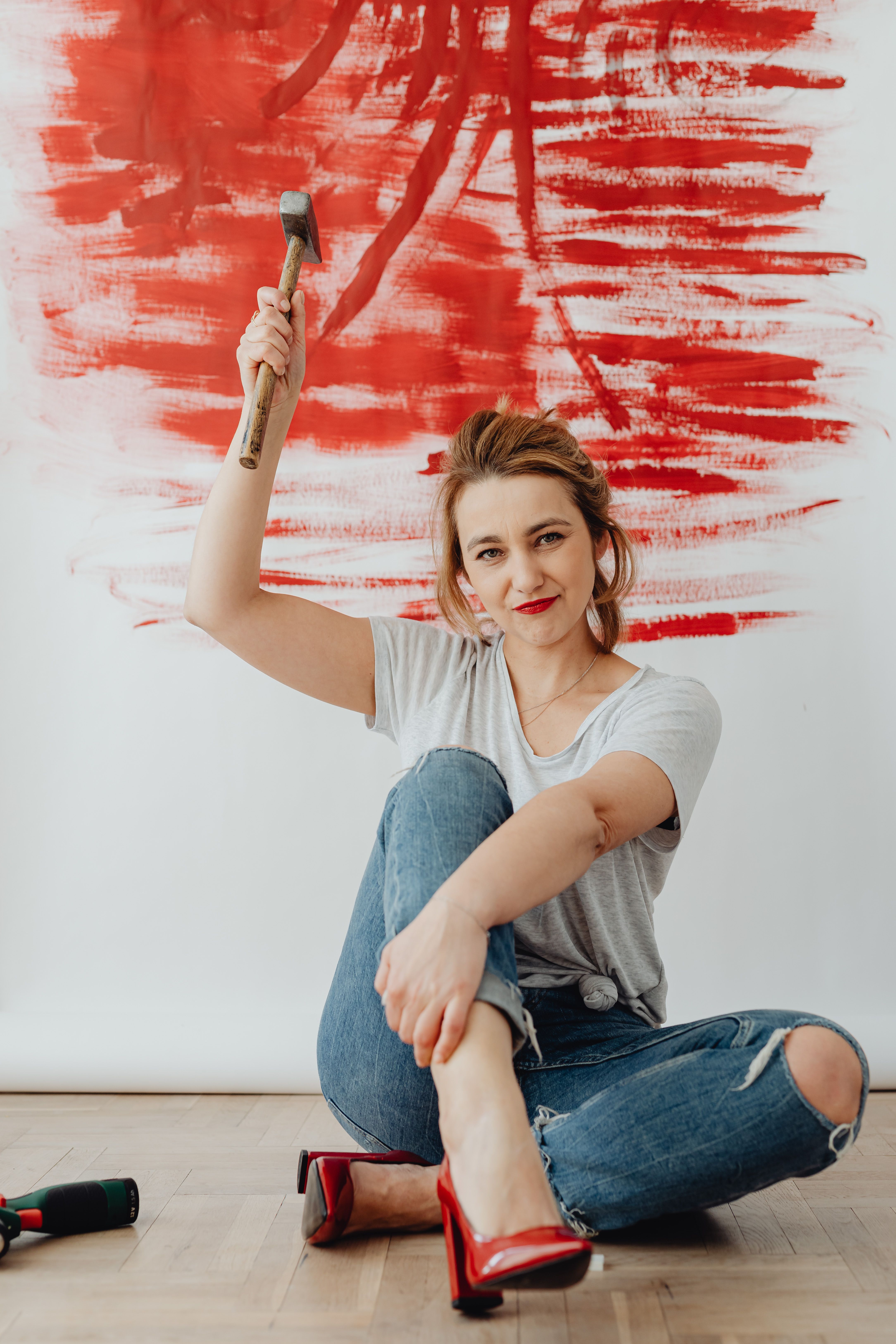 Woman sitting with a hammer in front of a red painted wall, smiling confidently.