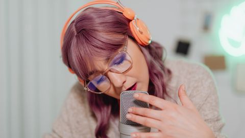 A young woman with vibrant hair and glasses recording audio with a microphone indoors.