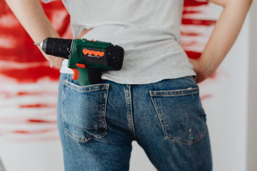 Close-up of a woman holding a power drill tucked into her denim pocket, with a red abstract background.