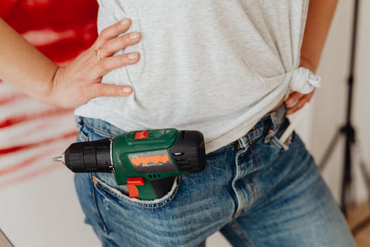 A close-up image of a person's hand on the waist with a power drill tucked into blue jeans.