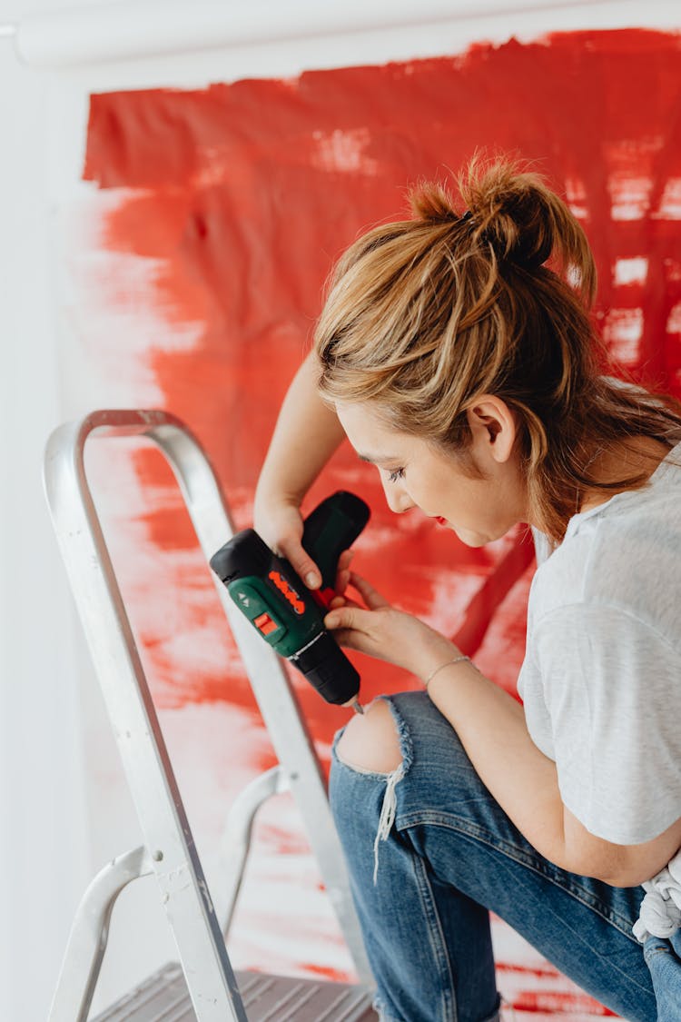 Woman With Hole In Jeans Drilling On A Ladder And Red Paint In Background