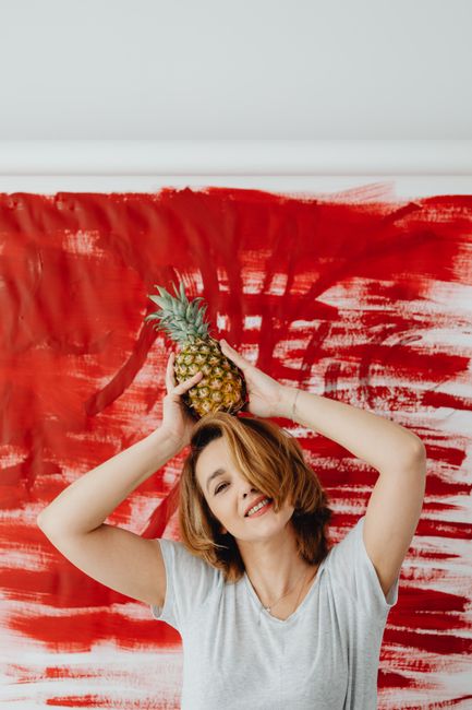Smiling woman poses with a pineapple, vibrant red backdrop, creative expression.
