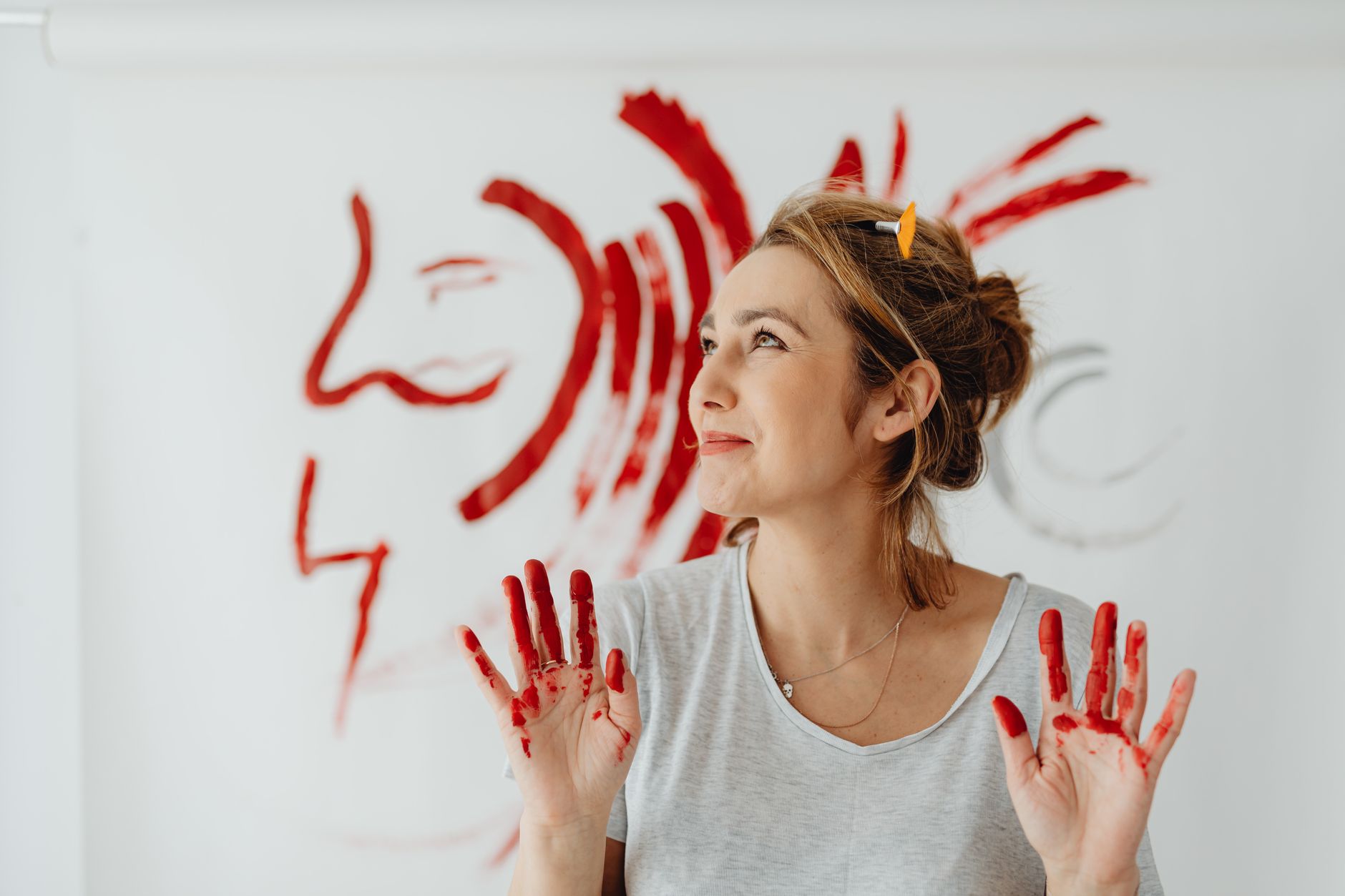 A woman smiles while showing her red paint-covered hands, standing in front of her abstract artwork.