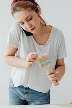 Caucasian woman multitasking with paintbrush and phone indoors.