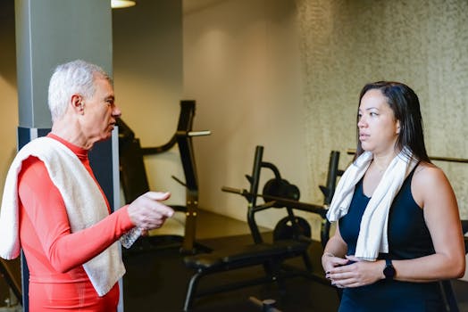 An elderly man and woman discuss fitness routines in a gym setting.