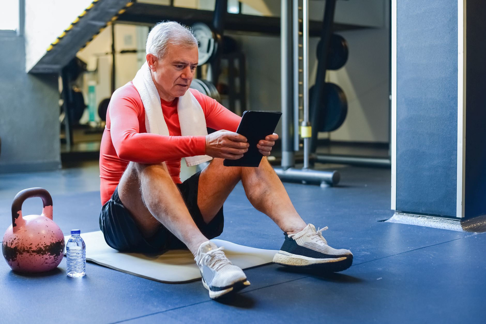 Elderly man in activewear using a tablet while resting on exercise mat in gym.