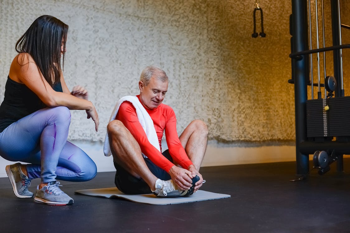 A Woman Training an Elderly Man Sitting on Yoga Mat 