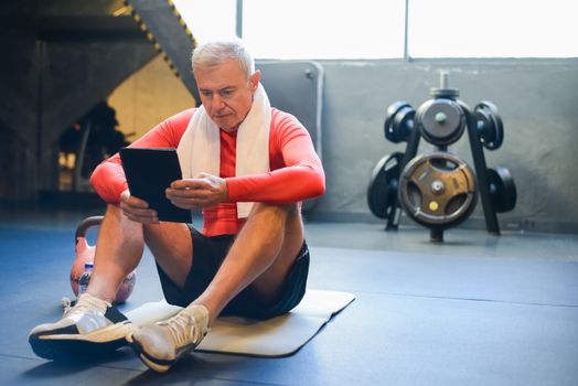 Elderly man in sportswear checking tablet during gym rest session.