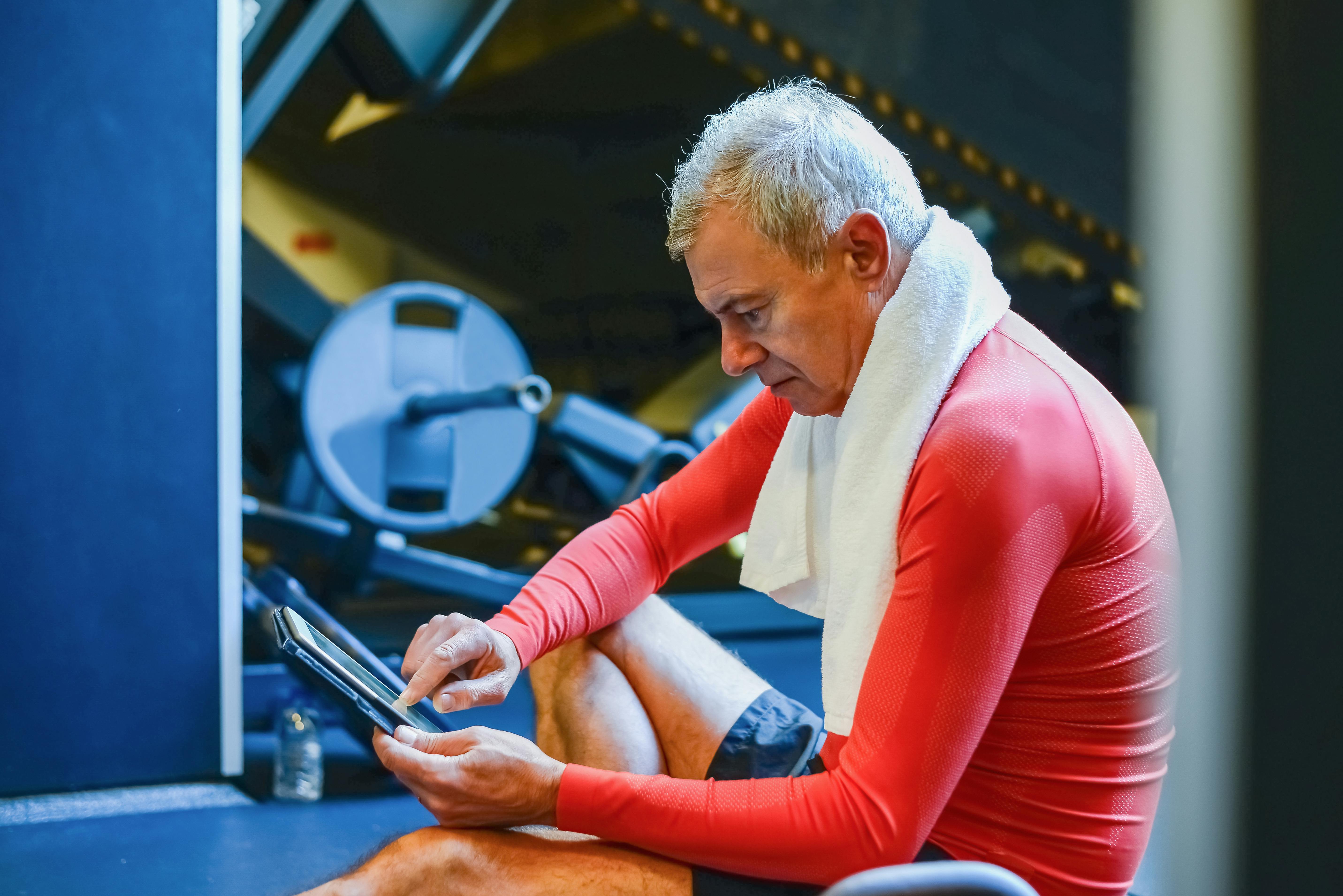 Senior man in red attire using a tablet in a gym environment. Fitness and technology blend.