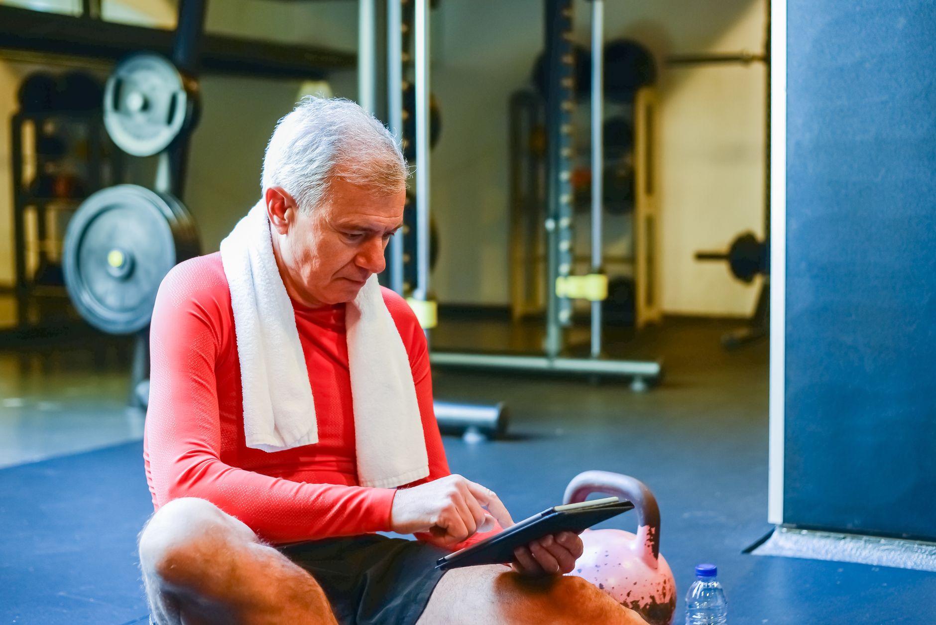Elderly man in gym with red top using tablet, showcasing active lifestyle and technology use.