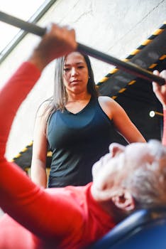 An elderly man performing bench press with a fitness coach supervising in the gym.