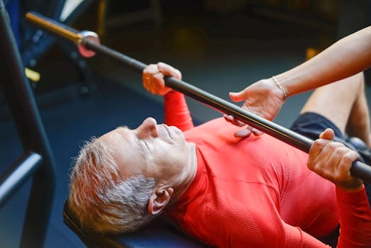 An elderly man performs a bench press exercise with assistance in a gym, promoting fitness and healthy living.