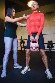 A senior man exercises with a kettlebell guided by a female trainer in a gym.