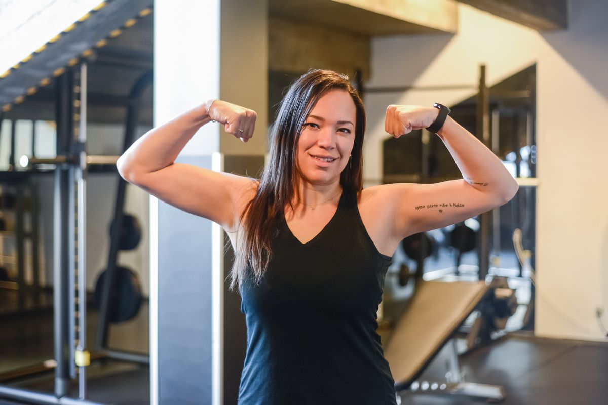 Una donna sorridente flette i bicipiti in palestra, esprimendo sicurezza e competenza