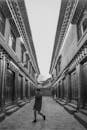 Grayscale Photo of A Person Walking on Street Between Buildings