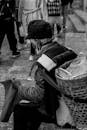 A Grayscale of an Elderly Woman Carrying a Basket on Her Back