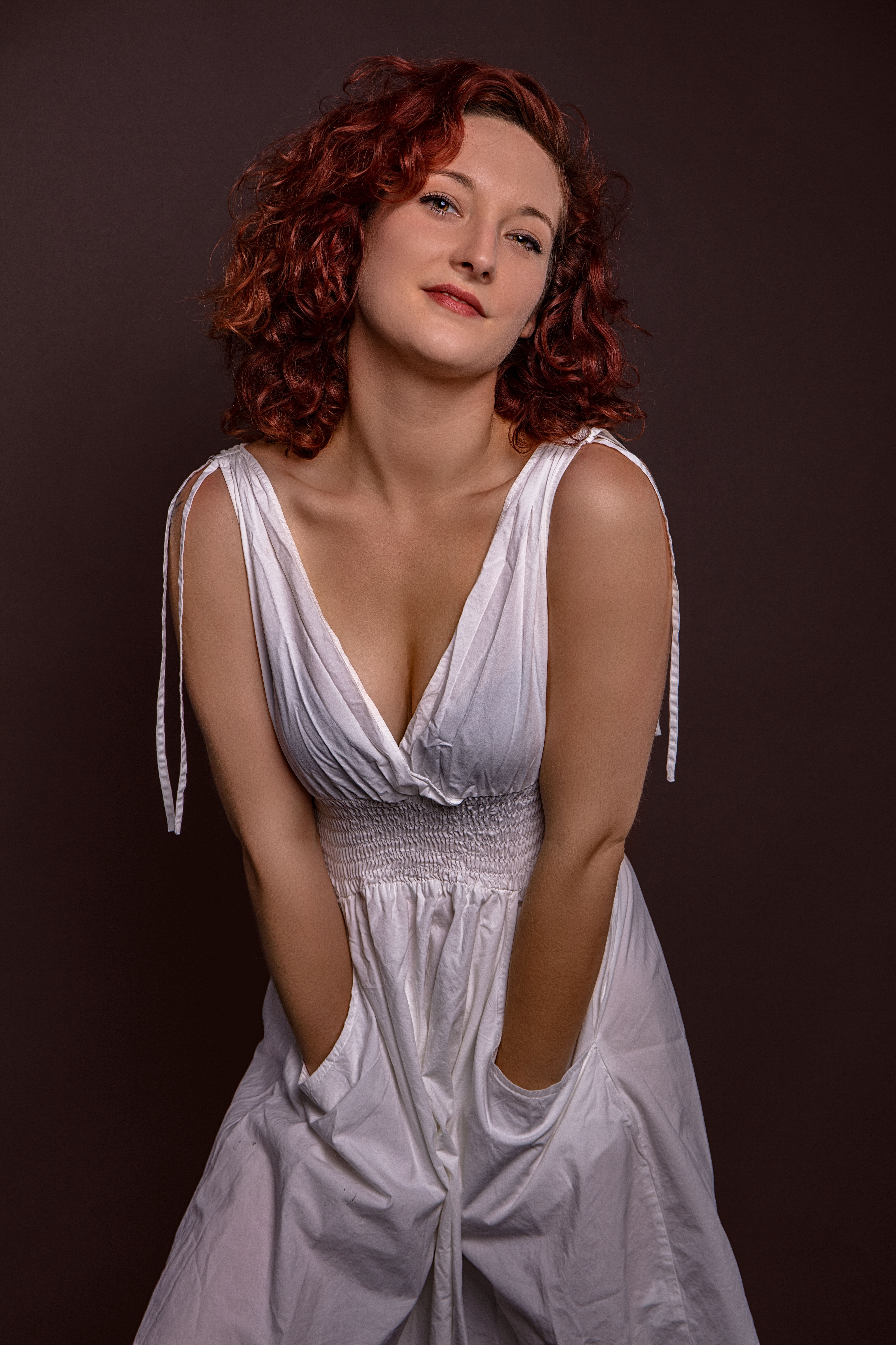 Elegant portrait of a woman posing with hands in pockets, wearing a white dress in a studio setting.
