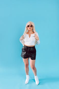 Fashionable woman wearing trendy white crop top, leather shorts, and sunglasses posing in studio against blue backdrop.