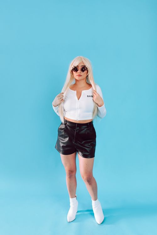 Free Fashionable woman wearing trendy white crop top, leather shorts, and sunglasses posing in studio against blue backdrop. Stock Photo