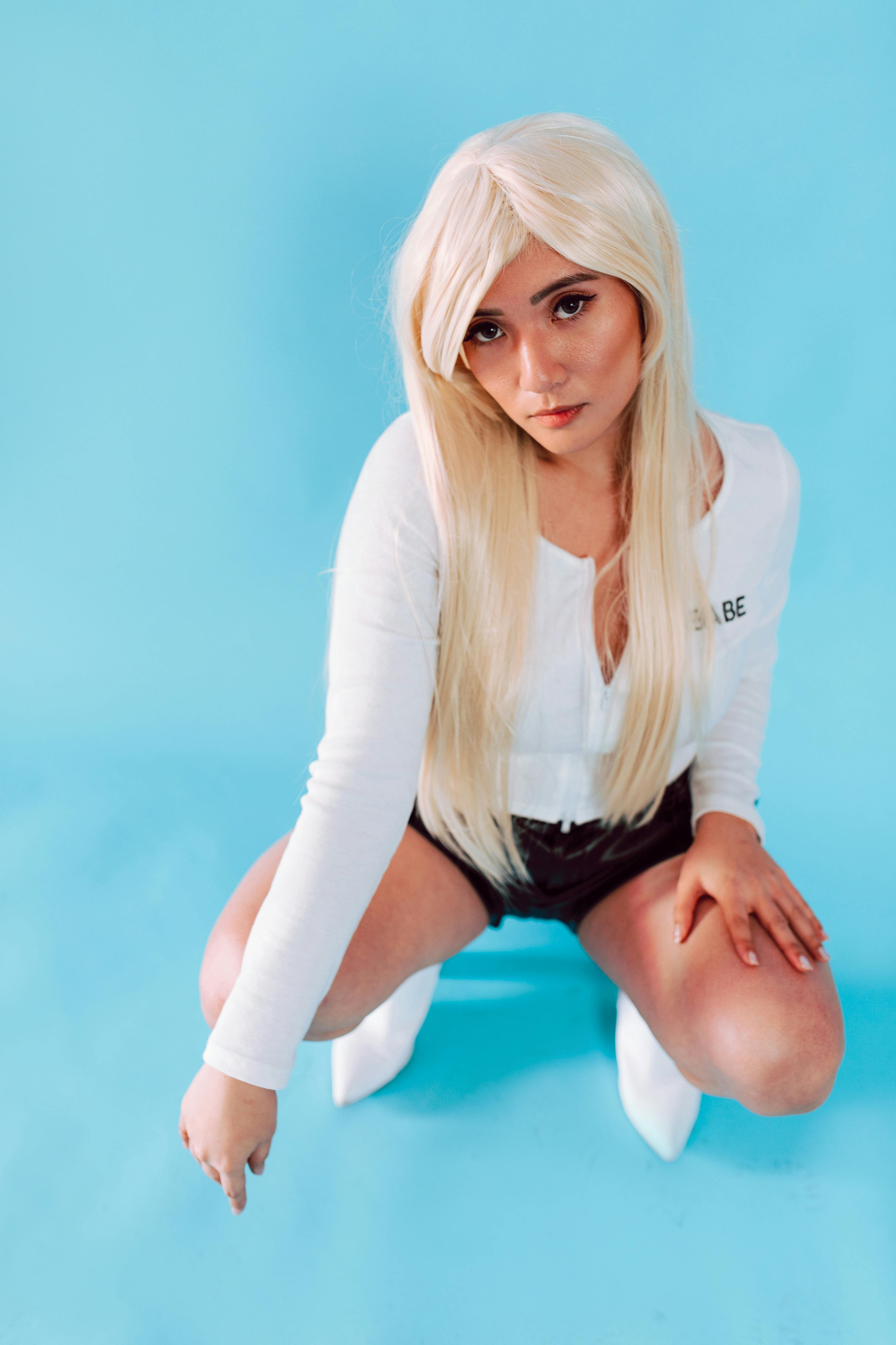 Stylish woman poses confidently in a blond wig and modern attire against a blue background.