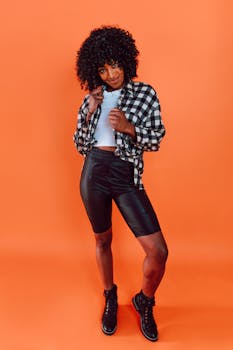 Stylish woman in leather shorts and plaid shirt against an orange background, radiating confidence.