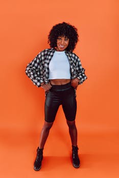 Confident woman with curly hair striking a pose in chic attire against a bright orange backdrop.