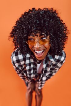 Vibrant portrait of a joyful woman with curly hair in sunglasses and checkered shirt against an orange backdrop.