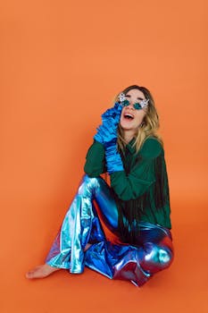 Colorful portrait of a woman in metallic pants and playful accessories against an orange backdrop.