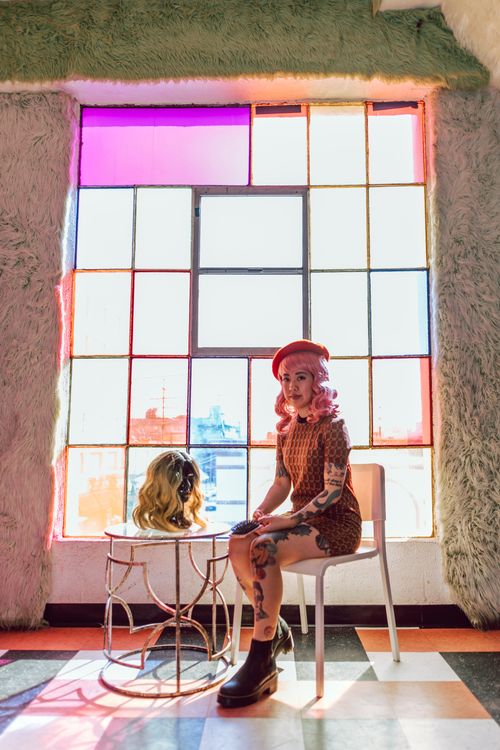 Free Stylish woman with colorful hair and tattoos seated indoors next to a wig on display. Vibrant atmosphere. Stock Photo