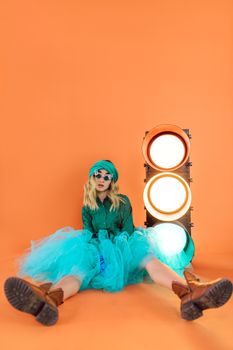 Stylish woman in teal dress poses with traffic light against vibrant orange backdrop.