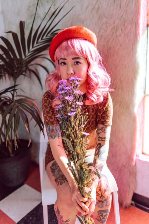 Free A fashionable woman with pink hair sitting indoors holding a bouquet of purple flowers. Stock Photo