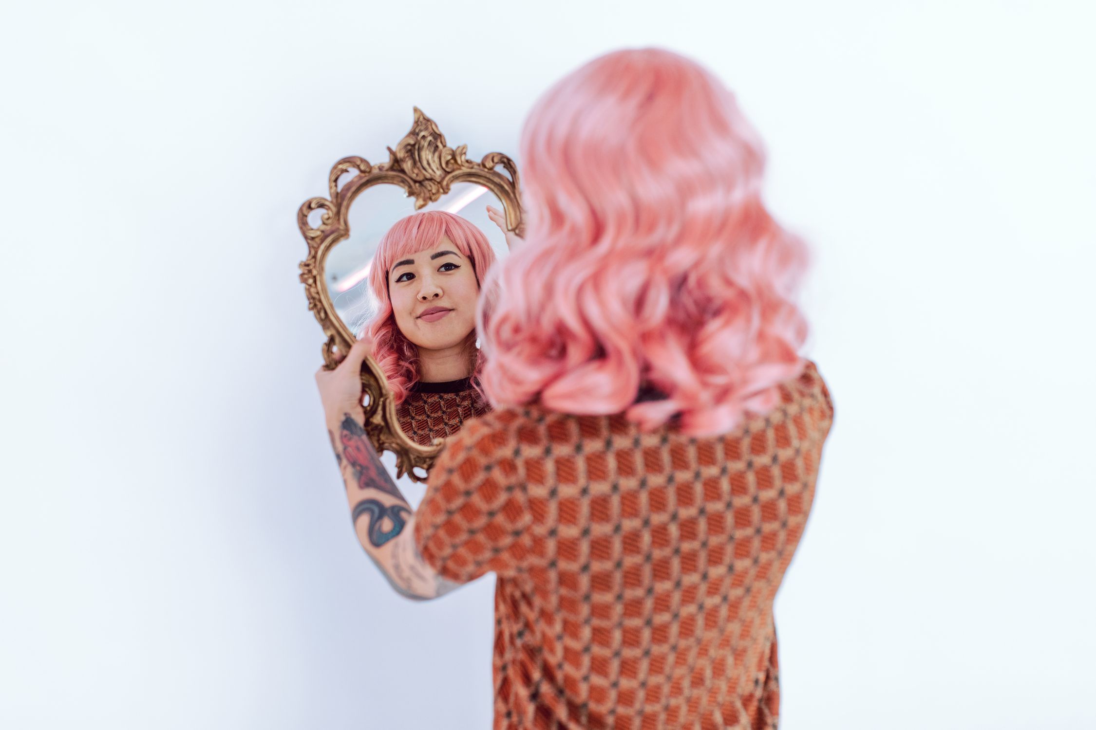 Mulher asiática com cabelo rosa e tatuagens admirando seu reflexo em um espelho decorado