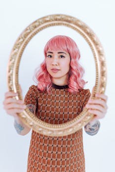 Woman holding ornate frame, showcasing pink hair and stylish pattern dress.
