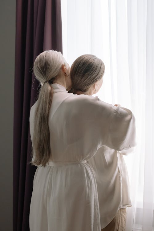 Free Women in White Long Sleeve Dress Standing Near the Window Stock Photo