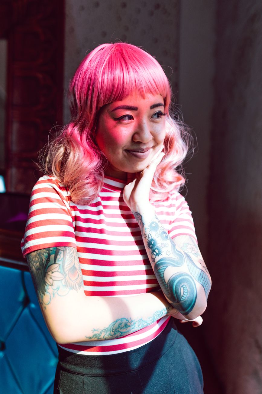 Stylish woman with pink hair and tattoos in a striped shirt, looking contemplatively to the side indoors.