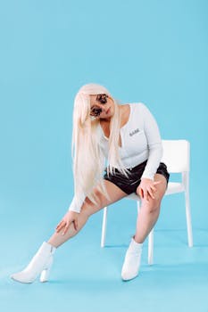 Fashionable woman wearing leather shorts and sunglasses poses on a chair against a blue background.