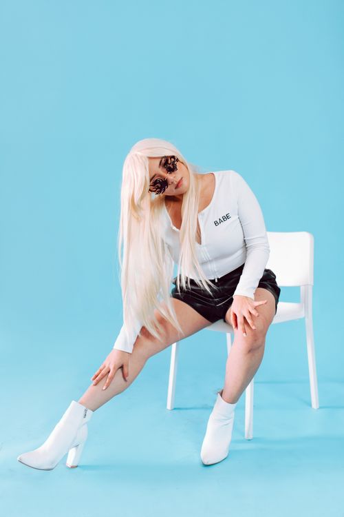 Free Fashionable woman wearing leather shorts and sunglasses poses on a chair against a blue background. Stock Photo