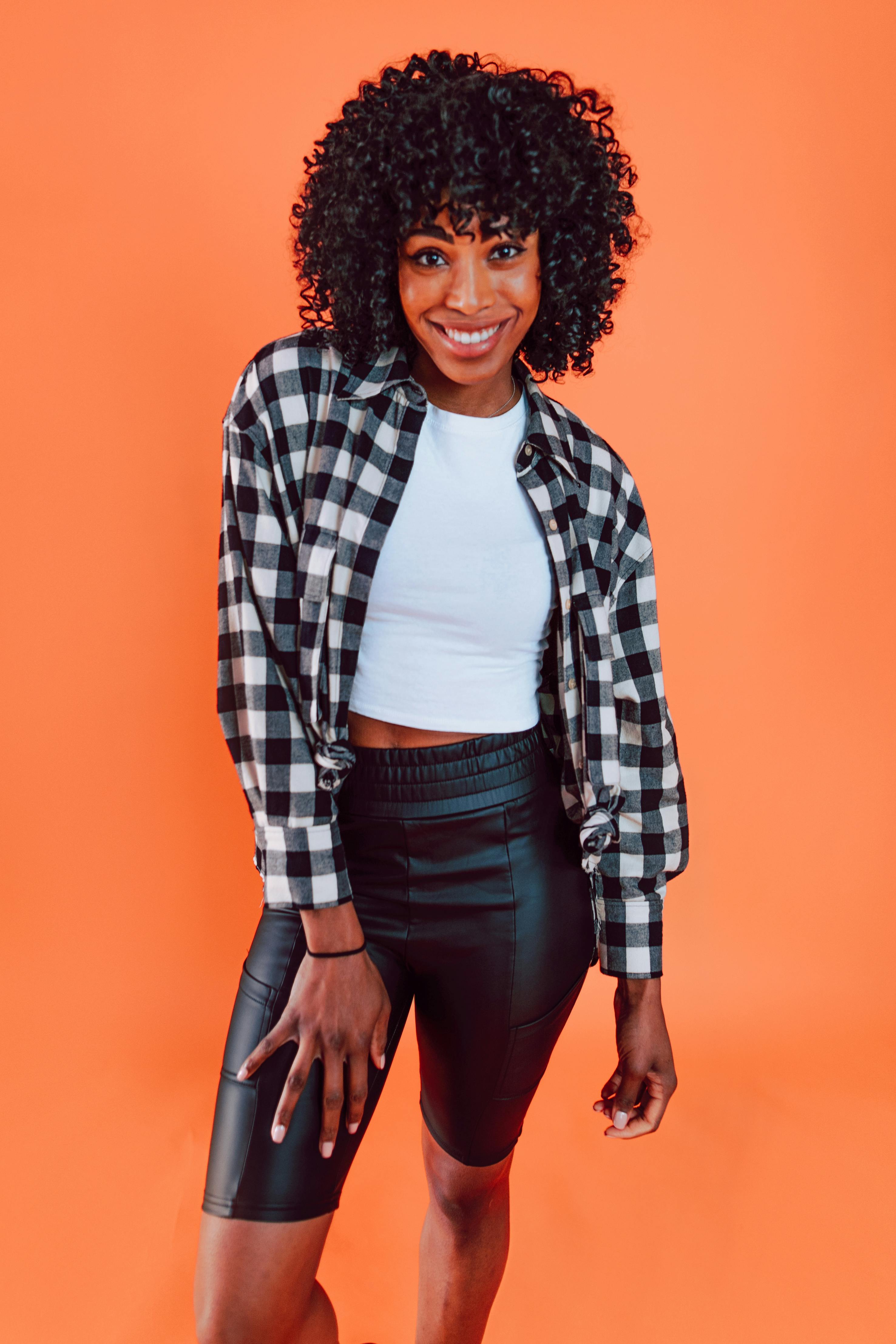 Portrait of a stylish woman wearing a plaid shirt and leather shorts against an orange backdrop.
