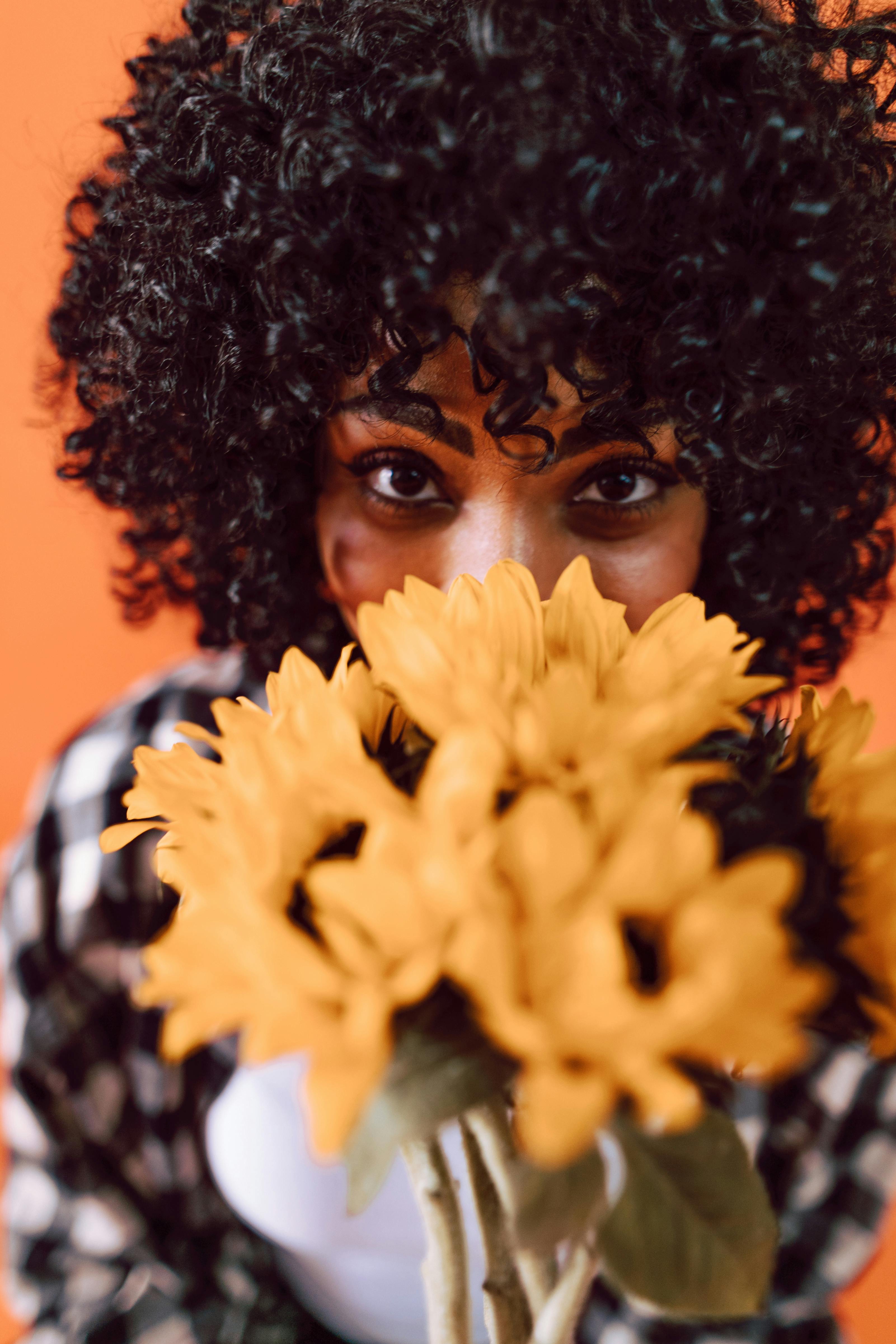 A captivating photo of a woman with curly hair holding sunflowers against an orange background.