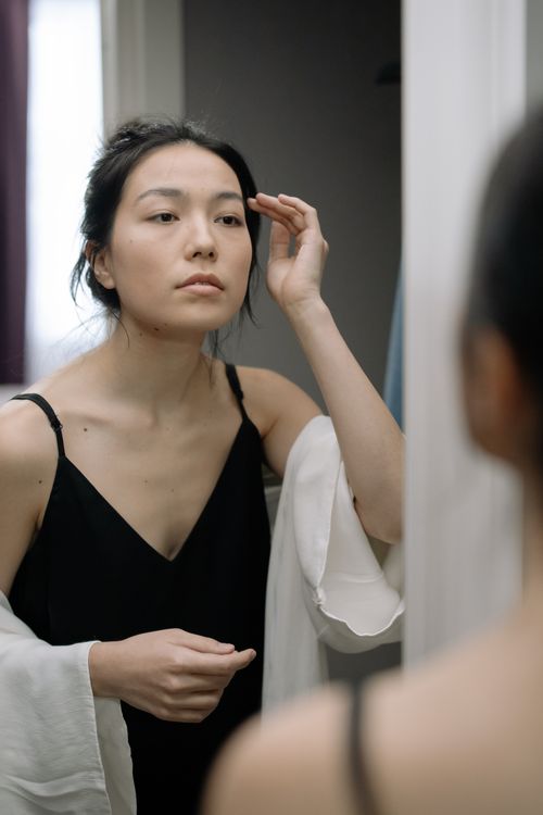 Free An Asian woman in a black nightdress examines her reflection in a mirror, contemplating her features. Stock Photo