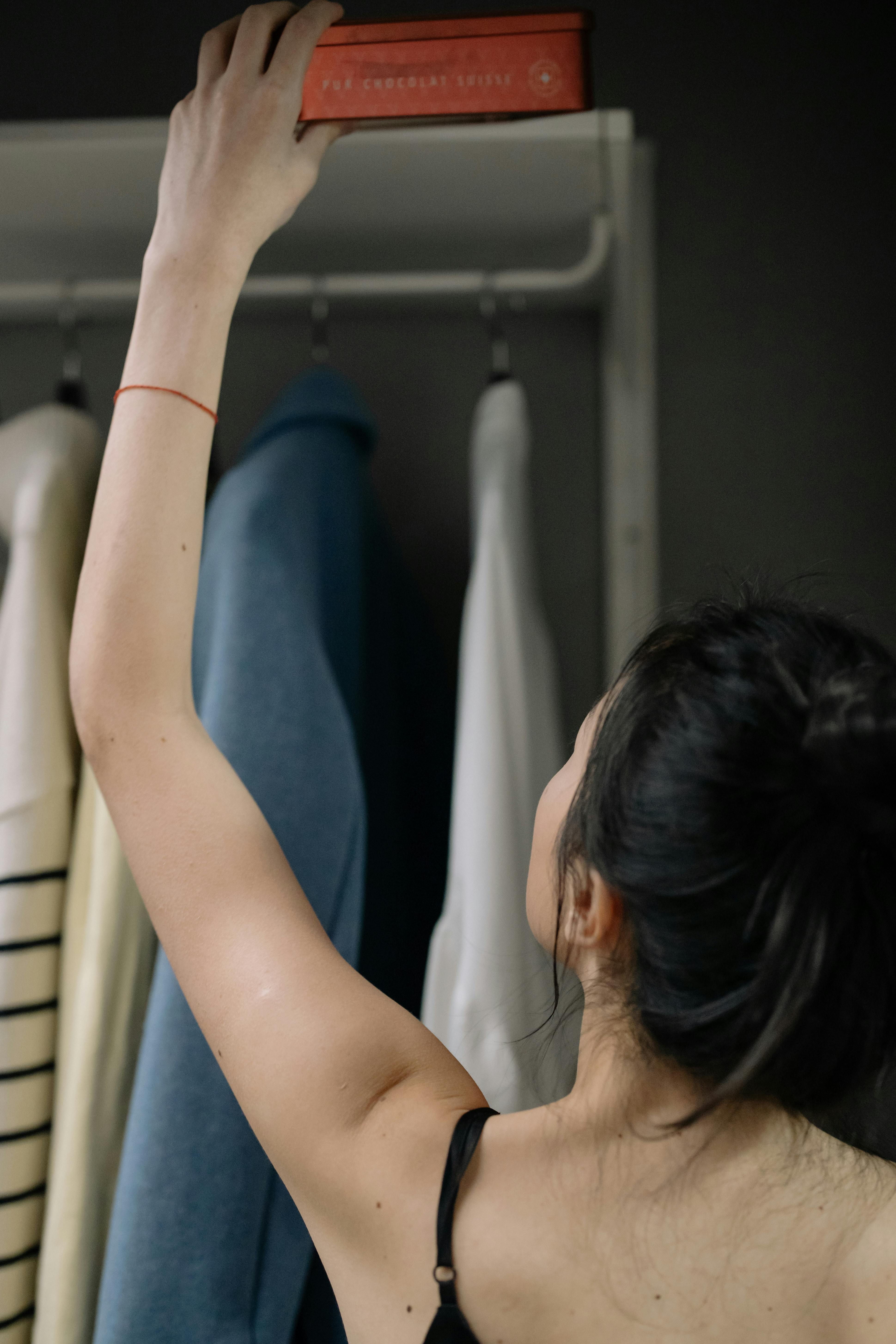 A woman in a spaghetti strap dress reaches for a red book in her closet.