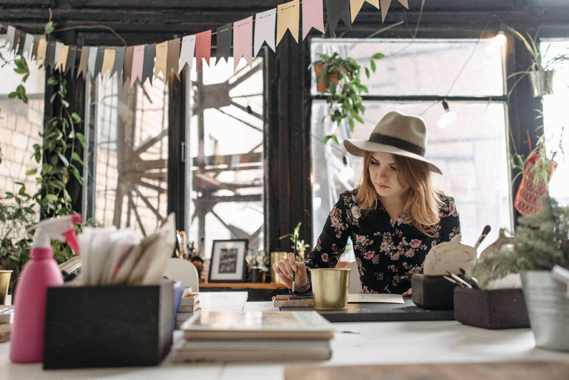 A young woman in a floral shirt and hat paints in a creative workshop filled with plants and natural light.