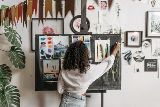 An artist with curly hair paints on a board surrounded by vibrant artworks and plants.