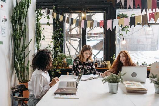 Three women working collaboratively in a stylish, plant-filled workspace featuring laptops, bunting, and natural light.